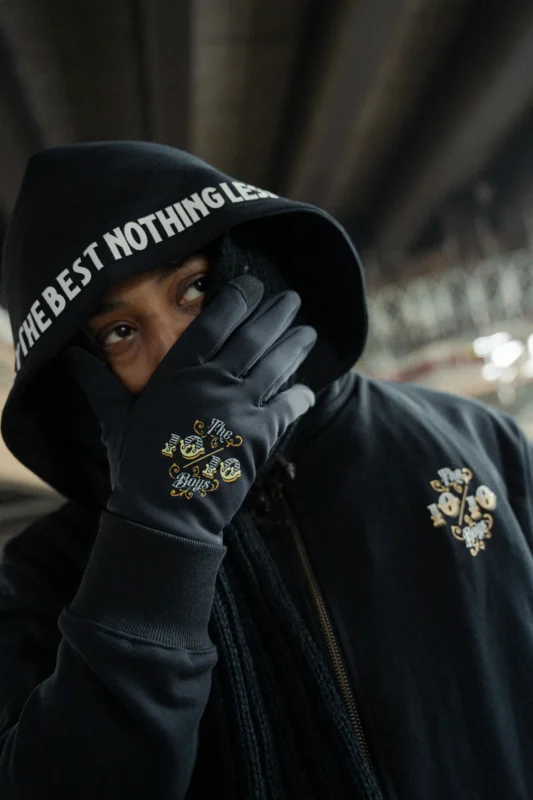 The 10/10 Boys Quality: model in a black hoodie with the hood up, highlighting clean stitching, embroidery, and a structured fit in a nighttime urban setting.