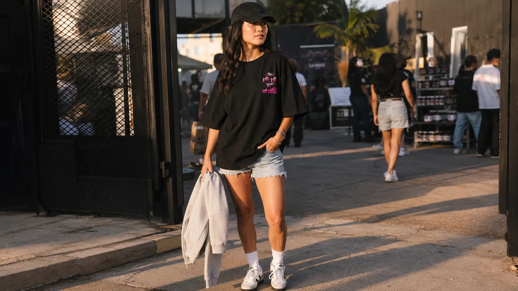 Day summer event outfit — oversized black tee with pink graphic, structured cap, crossbody bag and denim shorts, walking past a pop-up entrance