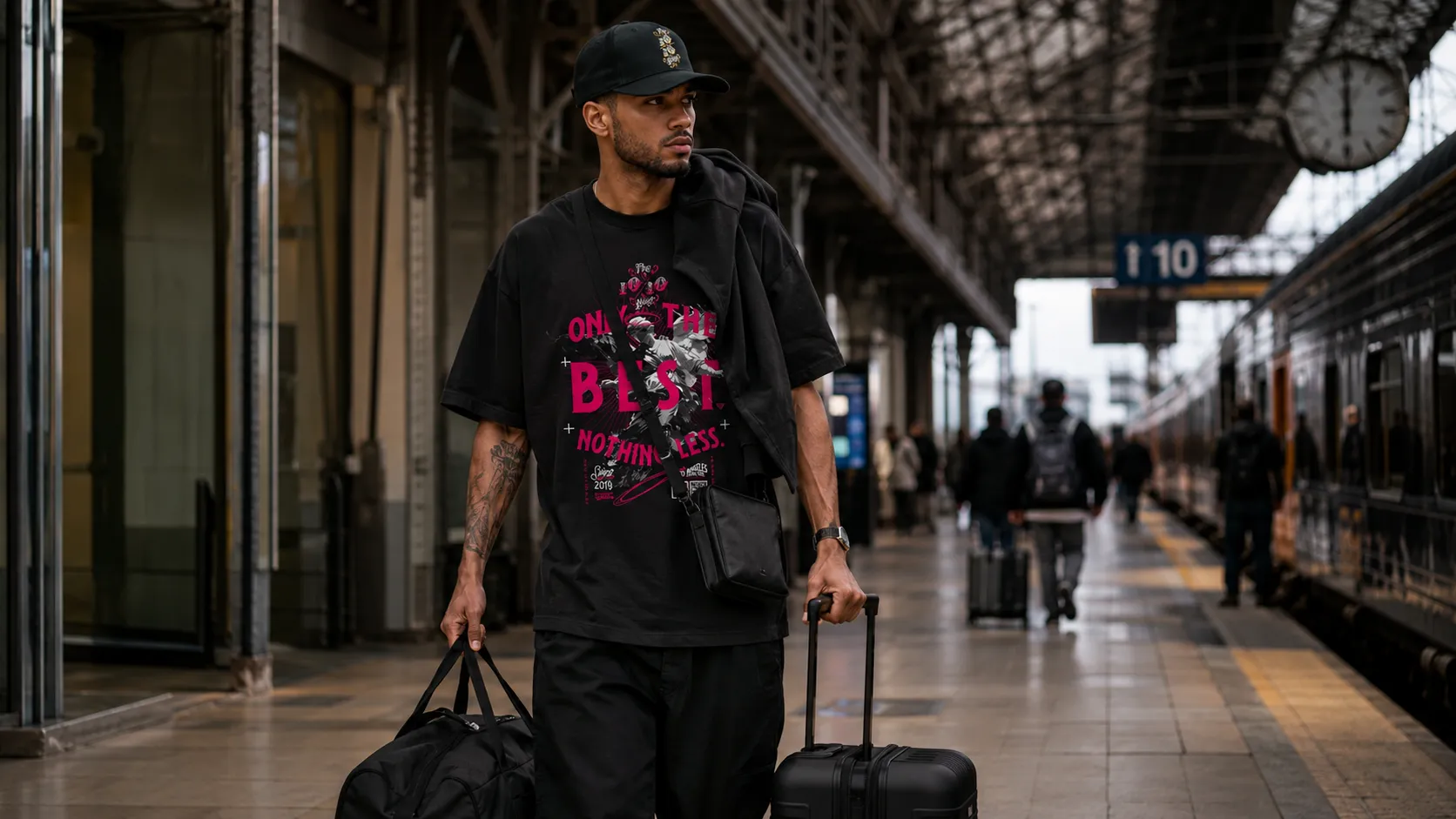 Day 1 travel fit in a black 10/10 Boys graphic tee and cap, carrying a duffel and suitcase on a train platform
