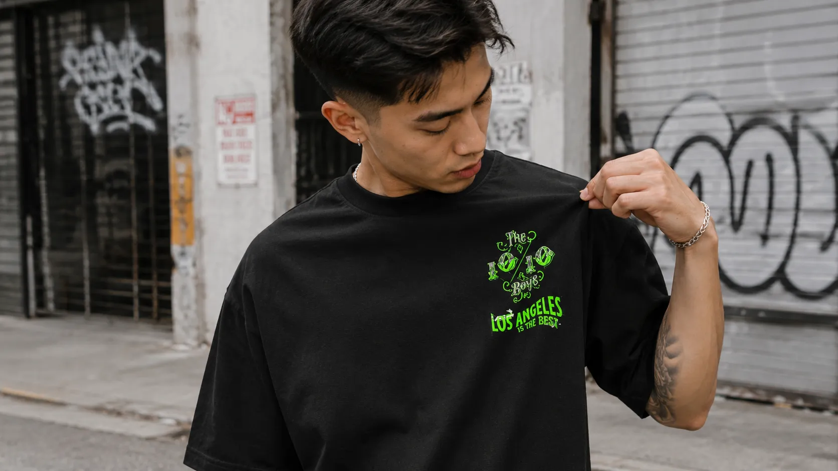 Close-up of a black oversized tee with a green 10/10 Boys Los Angeles chest print held up by the wearer