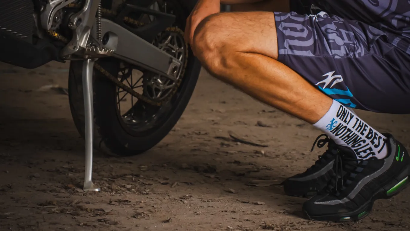 A person crouching beside a motorbike, the 10/10 Boys crew socks visible above black trainers