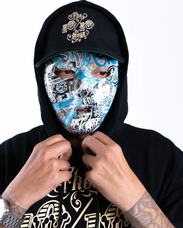 Masked figure with the hood up, wearing the 10/10 × GavinBrownArt mask with a 10/10 Boys cap and hoodie